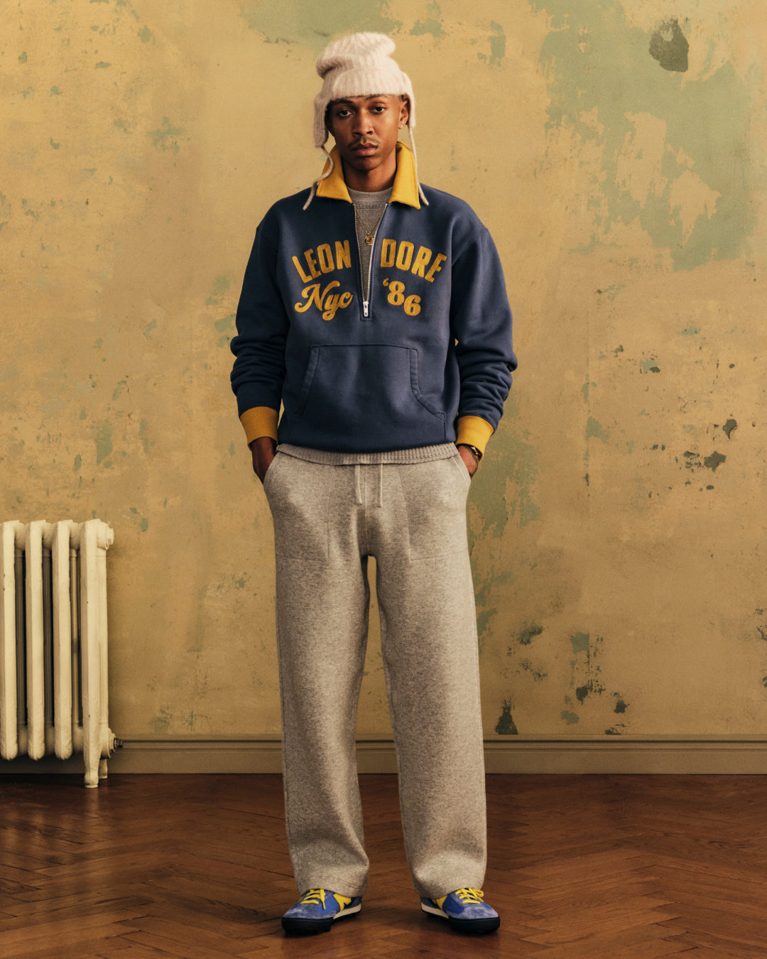 A person wearing an ALD / New Balance Gator, navy half-zip sweatshirt with yellow accents, grey trousers, and blue sneakers.