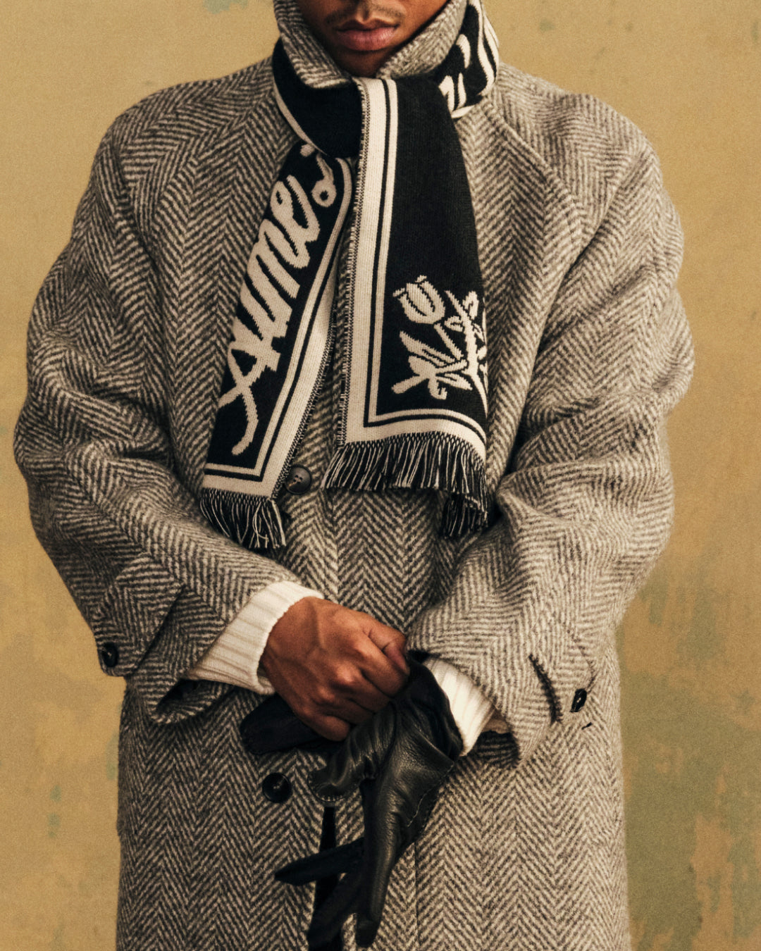 A person in a herringbone coat and patterned scarf holding black leather Henry Gloves.