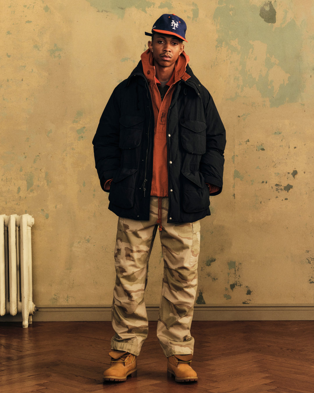 A man wears a black Down-Filled Surplus Parka over an orange sweatshirt with camouflage pants and a baseball cap.