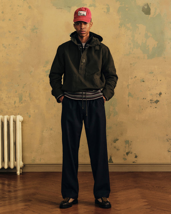 A person wears an Ottoman Stripe Quarter Zip Sweater over a striped knit, dark jacket, wide-leg trousers, and a red cap.