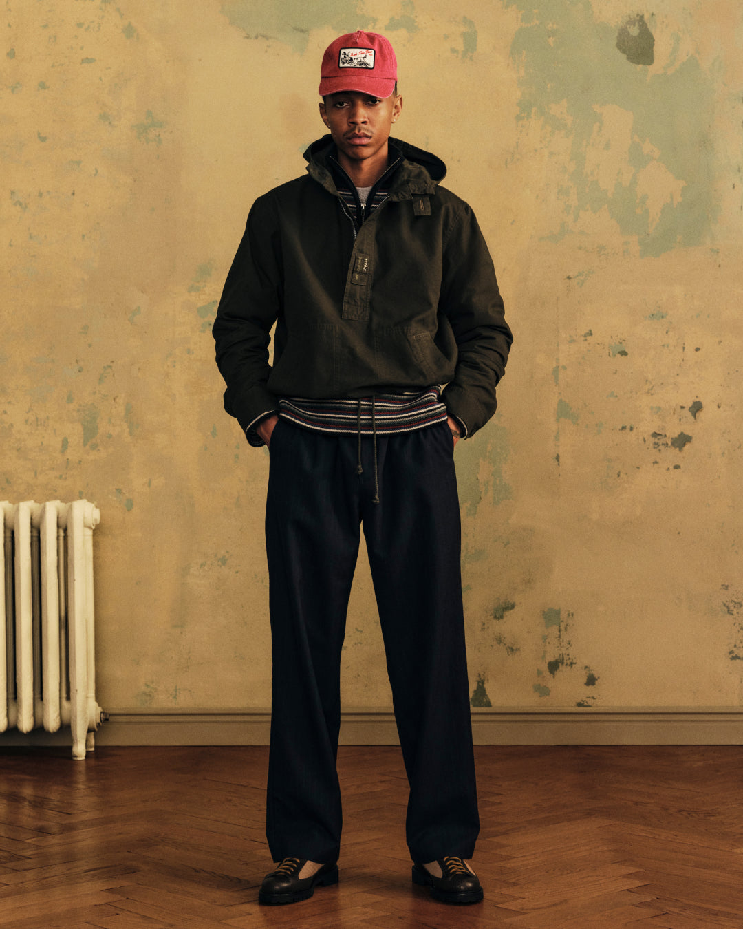 A person in a dark Garment Washed Anorak, red baseball cap, and wide-leg trousers stands against a weathered wall.