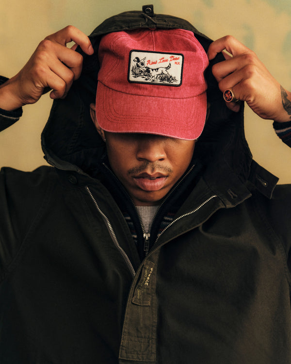 A person pulls the hood of a dark jacket over a red Aimé Retriever Hat featuring a graphic patch of a dog.