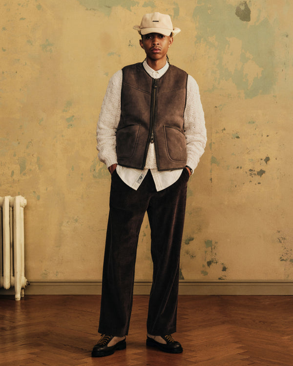 A man wearing a tan Shearling Earflap Hat, white textured shirt, brown zip-up vest, and dark corduroy trousers.