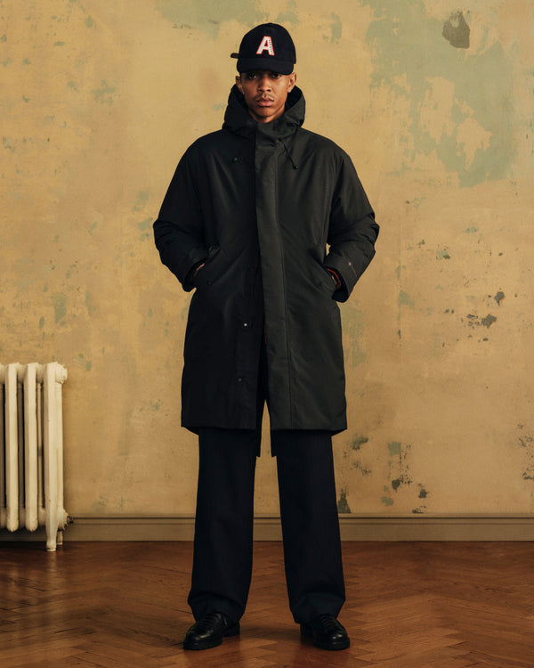 A person wearing a black GORE-TEX Down Filled Fishtail Parka, dark trousers, and an A baseball cap.