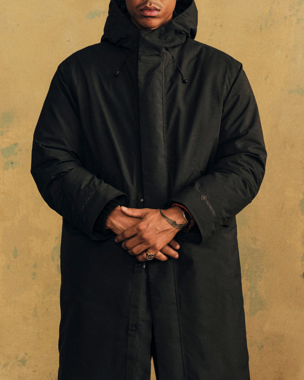 A person wearing a black GORE-TEX Down Filled Fishtail Parka with hands clasped in front.