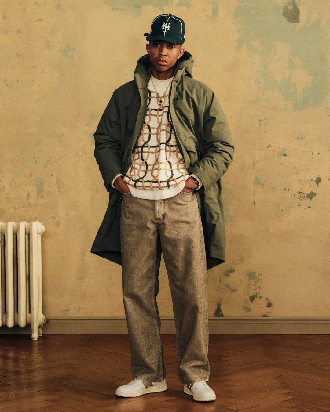 A man wears a cream Cable Link Crewneck Sweater, a green hooded parka, brown jeans, and a baseball cap.