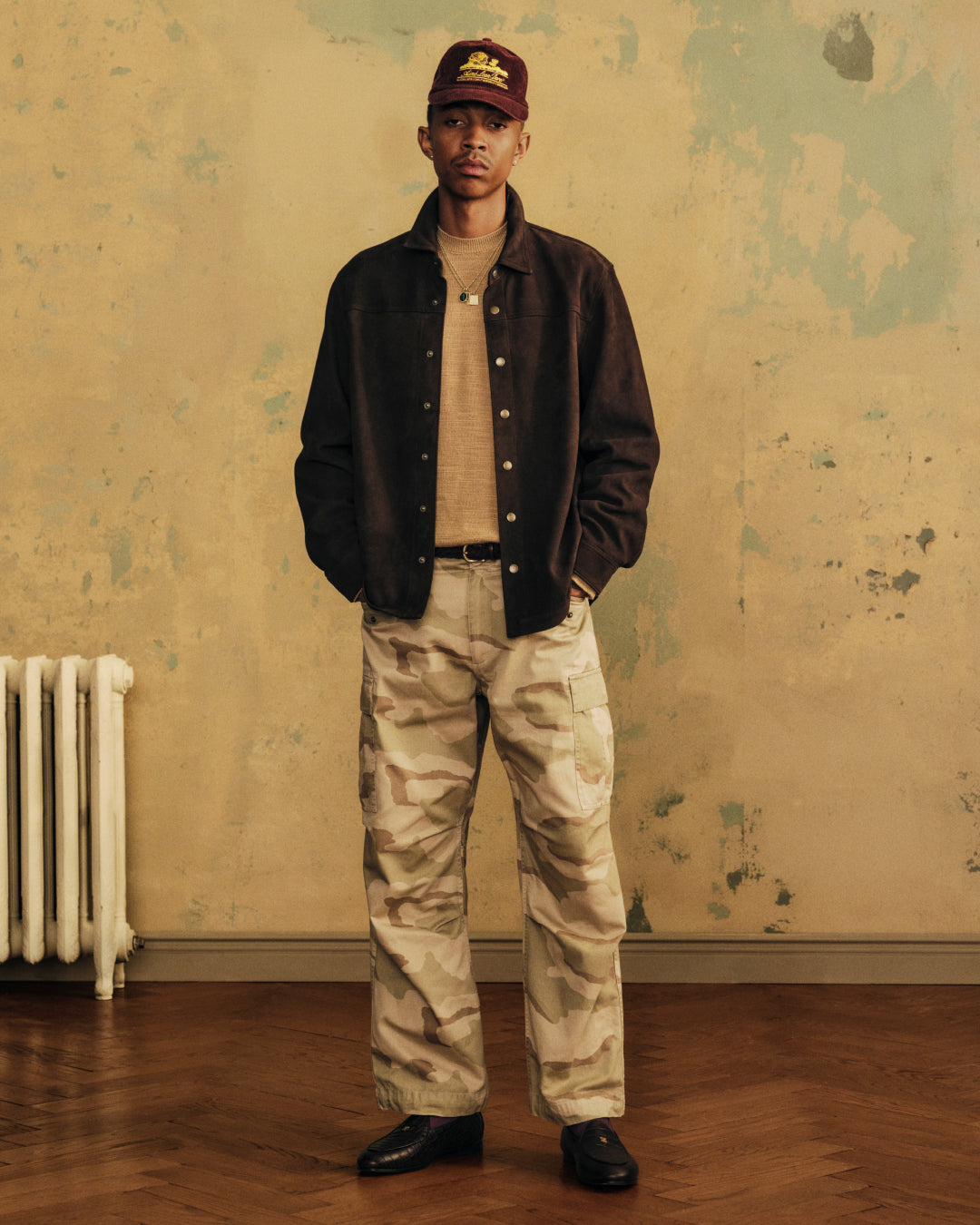 A person wears a brown Suede Overshirt over a tan sweater, camouflage cargo pants, and a maroon cap.