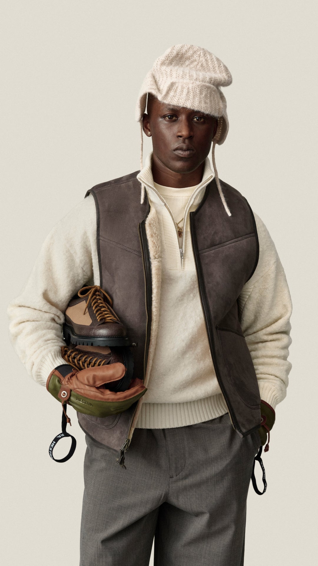 A model wearing a cream knit beanie with ear flaps, a brown leather vest over a cream sweater, gray trousers, and holding a brown leather boot and glove, set against a solid light beige background.
