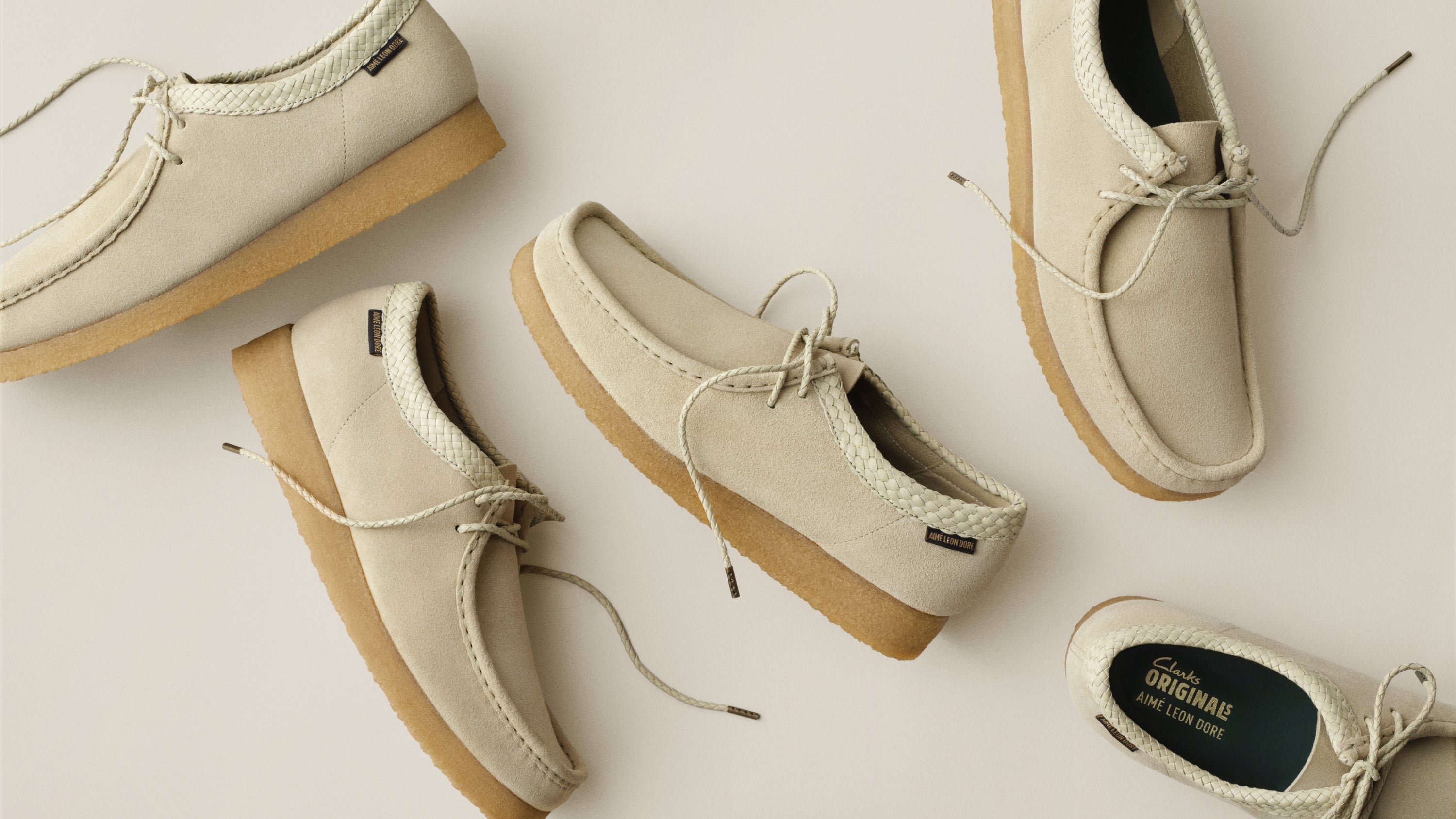 Five cream-colored suede slip-on shoes with tan soles and braided details, arranged in a flat-lay pattern on a light beige background. One shoe is shown from the top, one from the side, and one from the bottom revealing the 'The Wanderers' brand label.