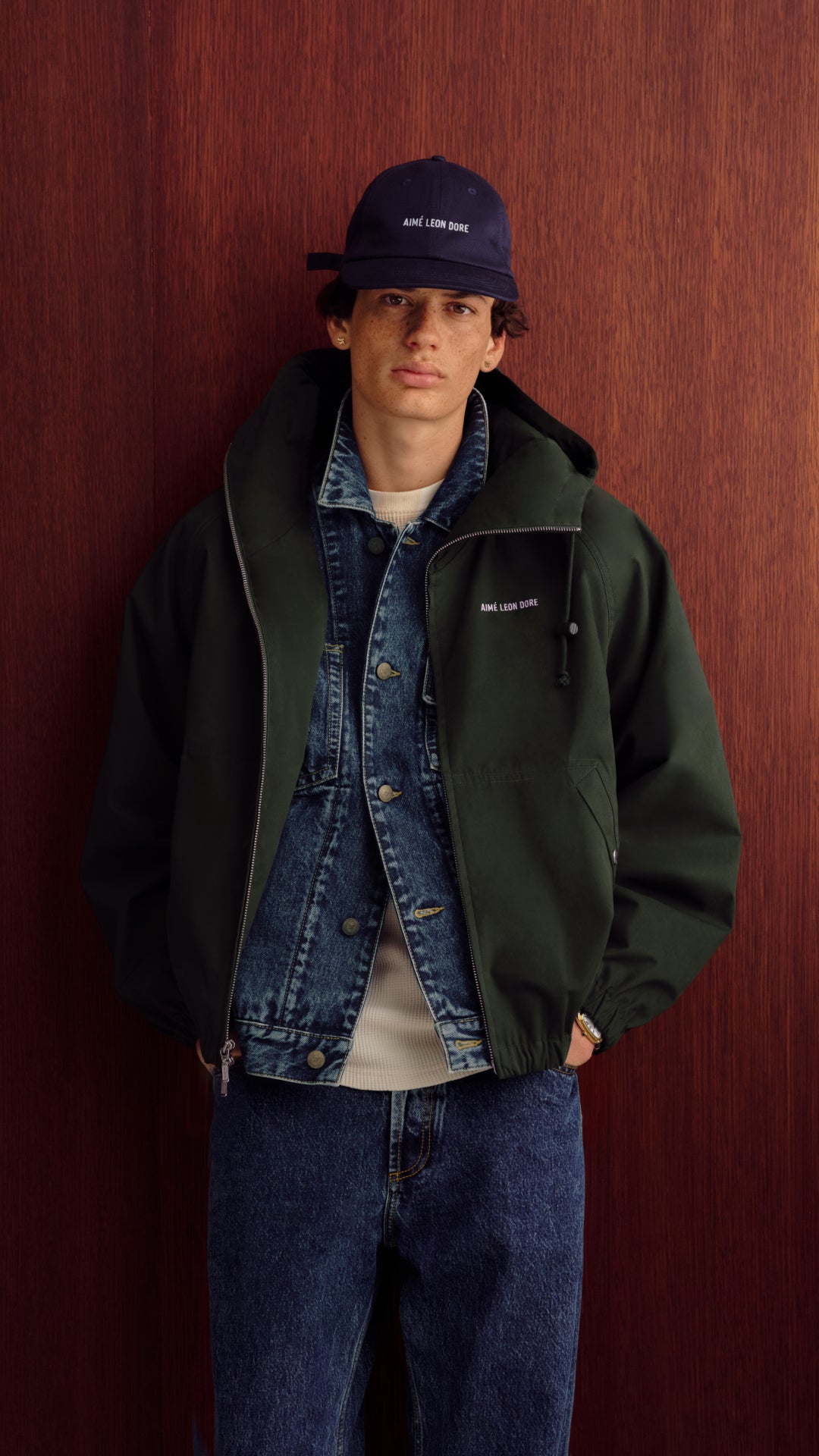 A man wearing a dark green hooded jacket with a small white embroidered logo on the chest, layered over a denim jacket and t-shirt, paired with a navy blue baseball cap, standing against a dark wood-paneled wall.