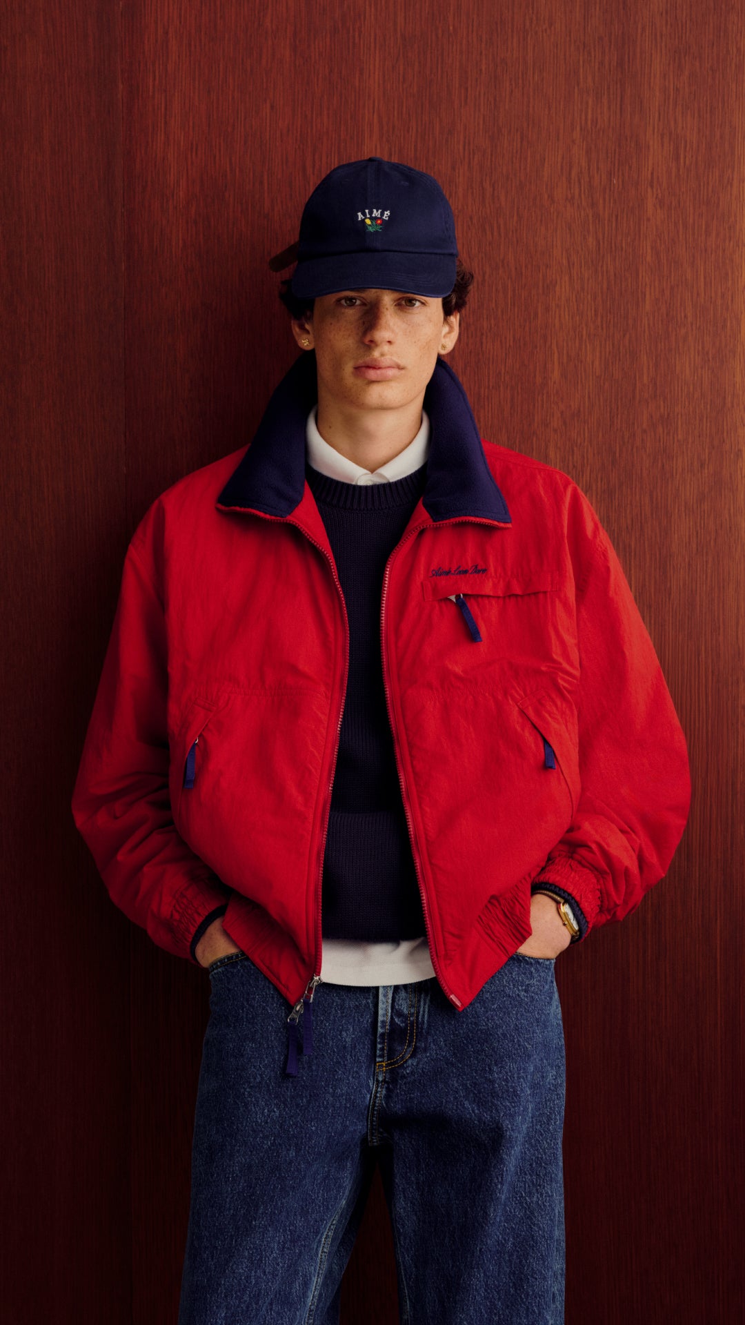 A model stands against a dark wood-paneled wall wearing a bright red zip-up jacket with a navy blue collar, a navy blue knit sweater, blue jeans, and a navy blue baseball cap.