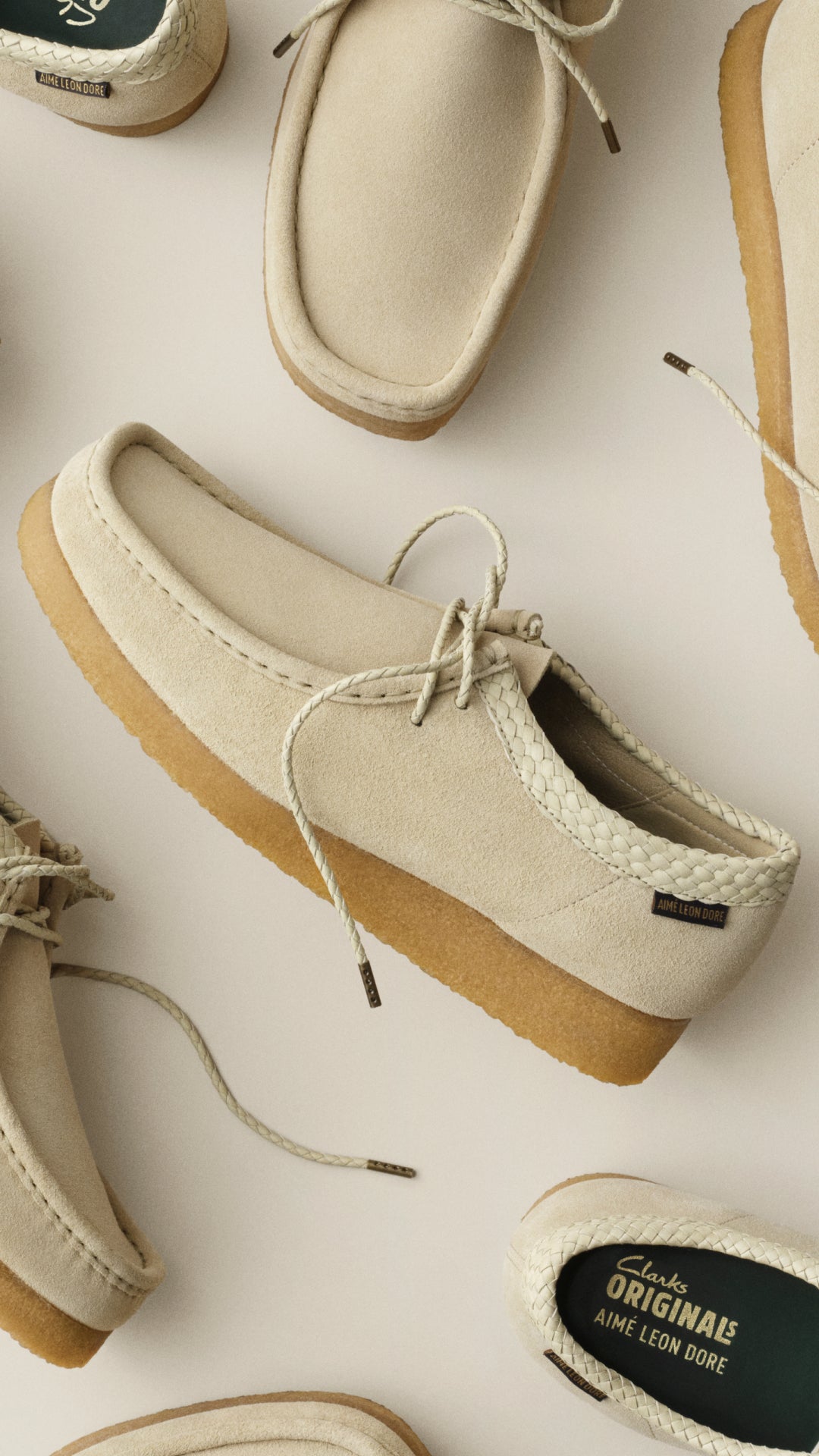 A flat-lay view of multiple cream suede lace-up shoes arranged on a light beige background, with one shoe prominently shown from the side and top. The shoes have tan soles and a small black brand tag on the side.