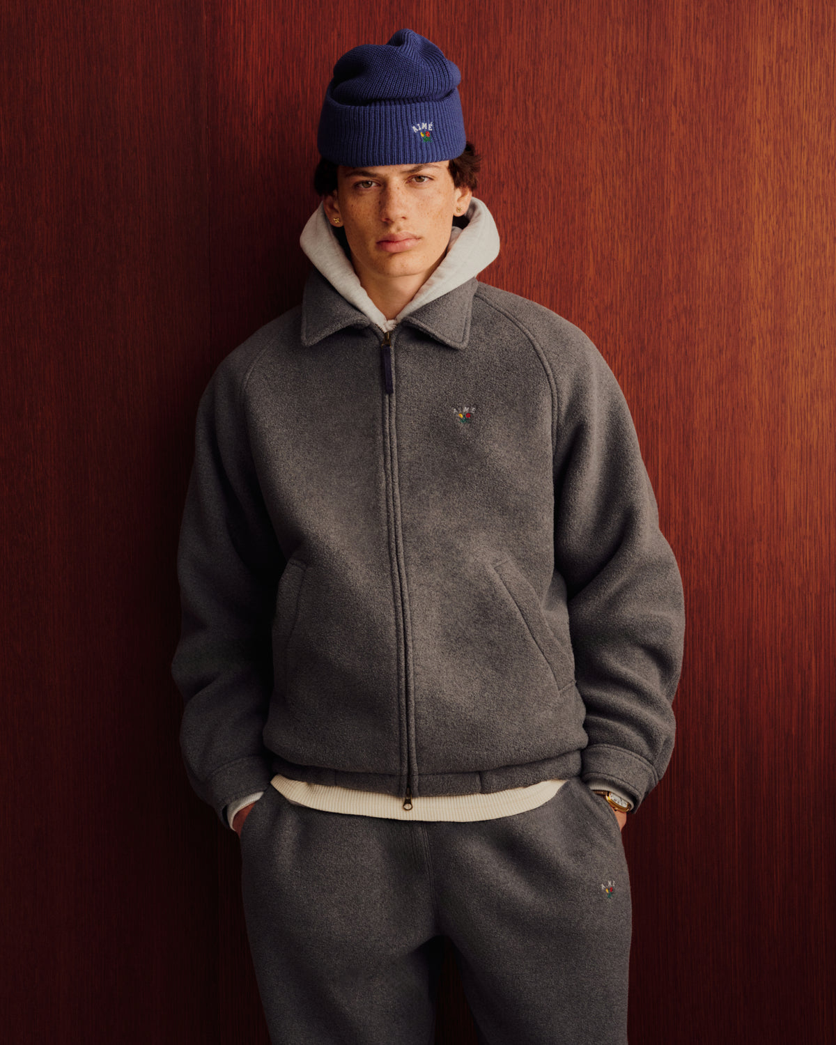Someone wears the Crest Fleece Casual Jacket in gray and poses against a wooden wall, styled with matching pants.