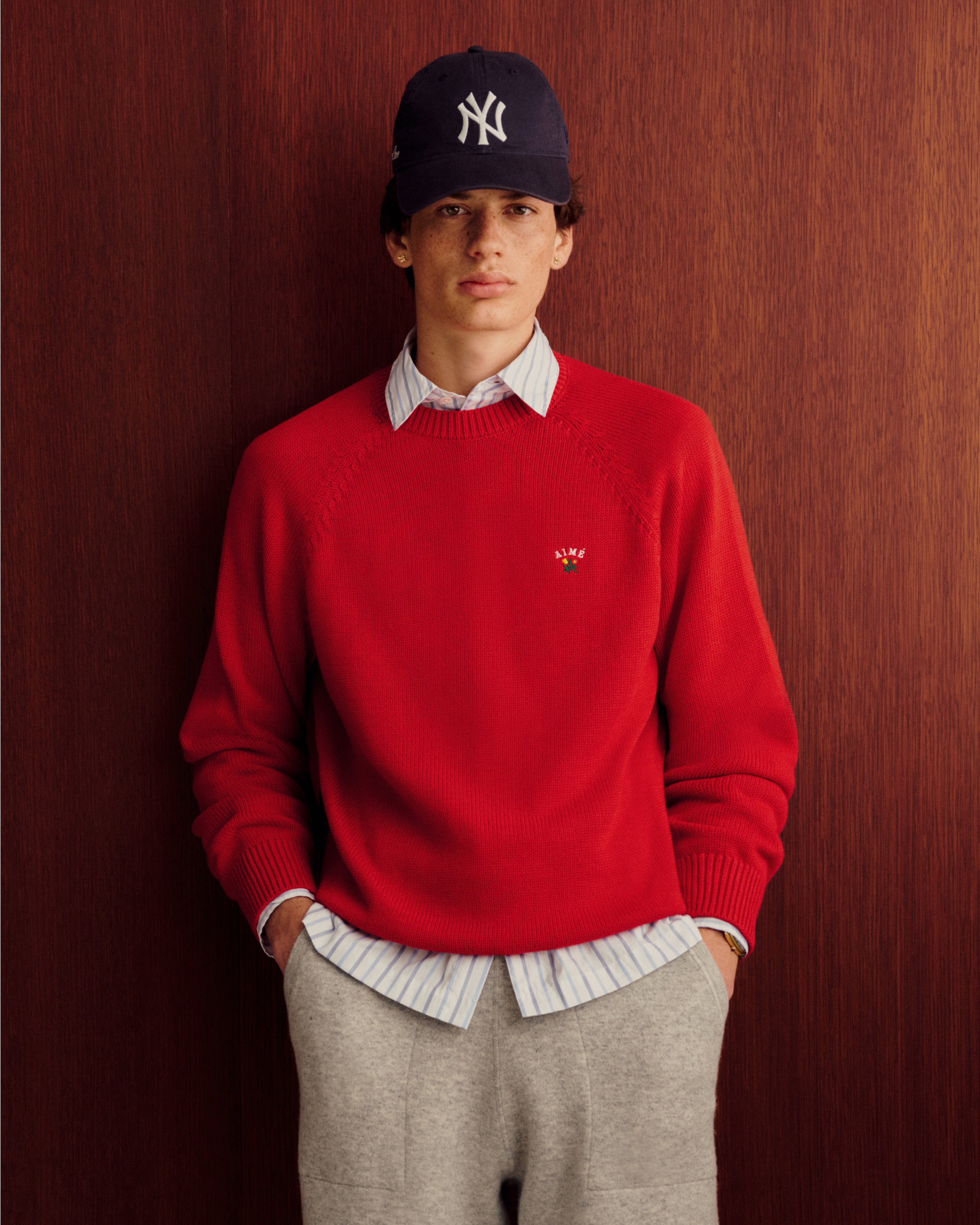A person in Pavilion Cashmere Sweatpants, a red sweater, and navy NY cap stands by a wooden background, hands in pockets.