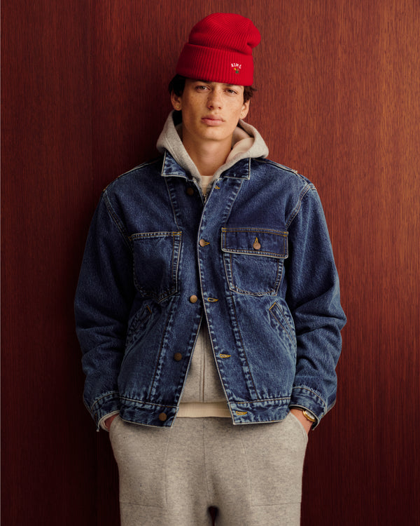 Young man in a red beanie, denim jacket, and Full-Zip Pavilion Cashmere Hoodie stands by a wood-paneled wall.