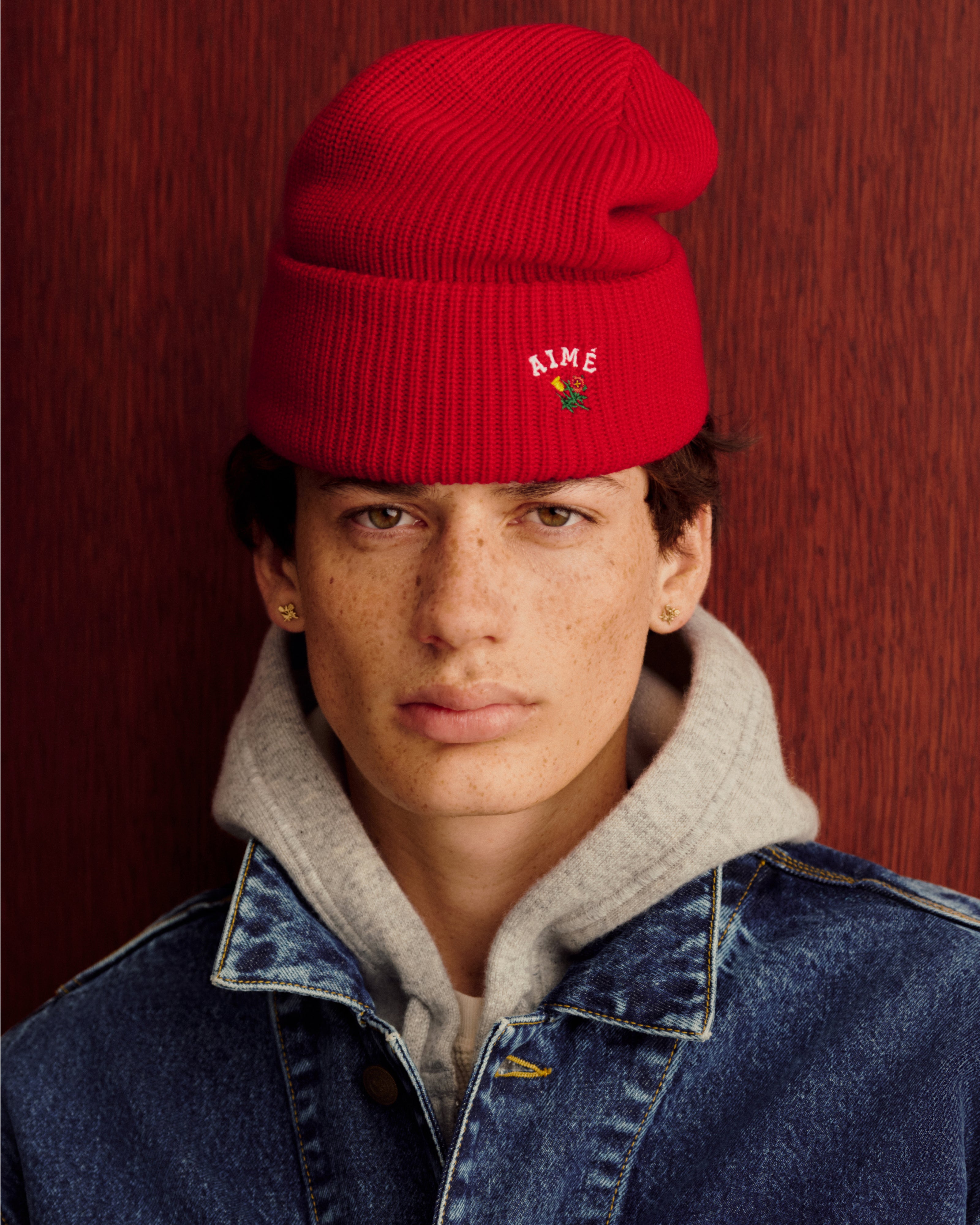 Young person in a Crest Merino Wool Beanie, gray hoodie, and denim jacket by a reddish wood wall.