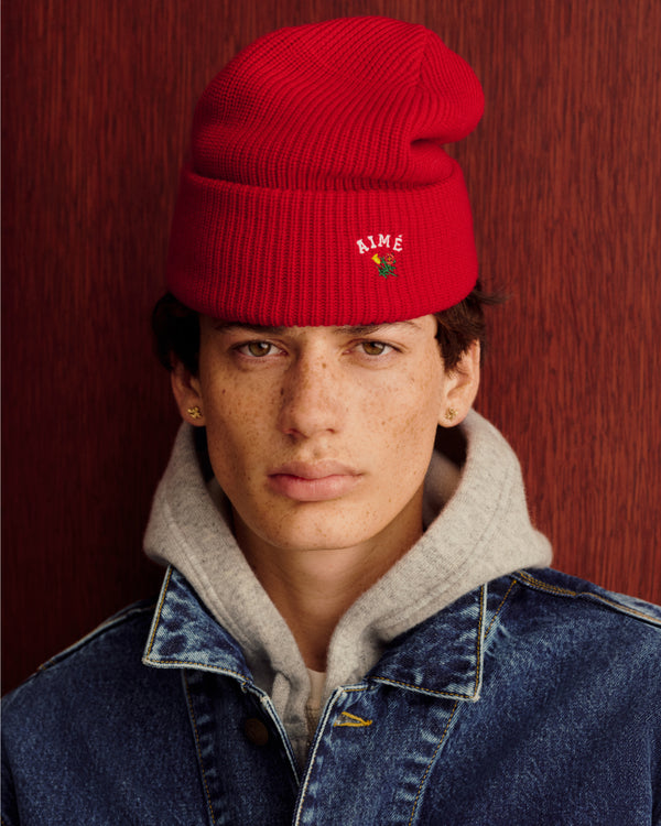 Young person in a Crest Merino Wool Beanie, gray hoodie, and denim jacket by a reddish wood wall.