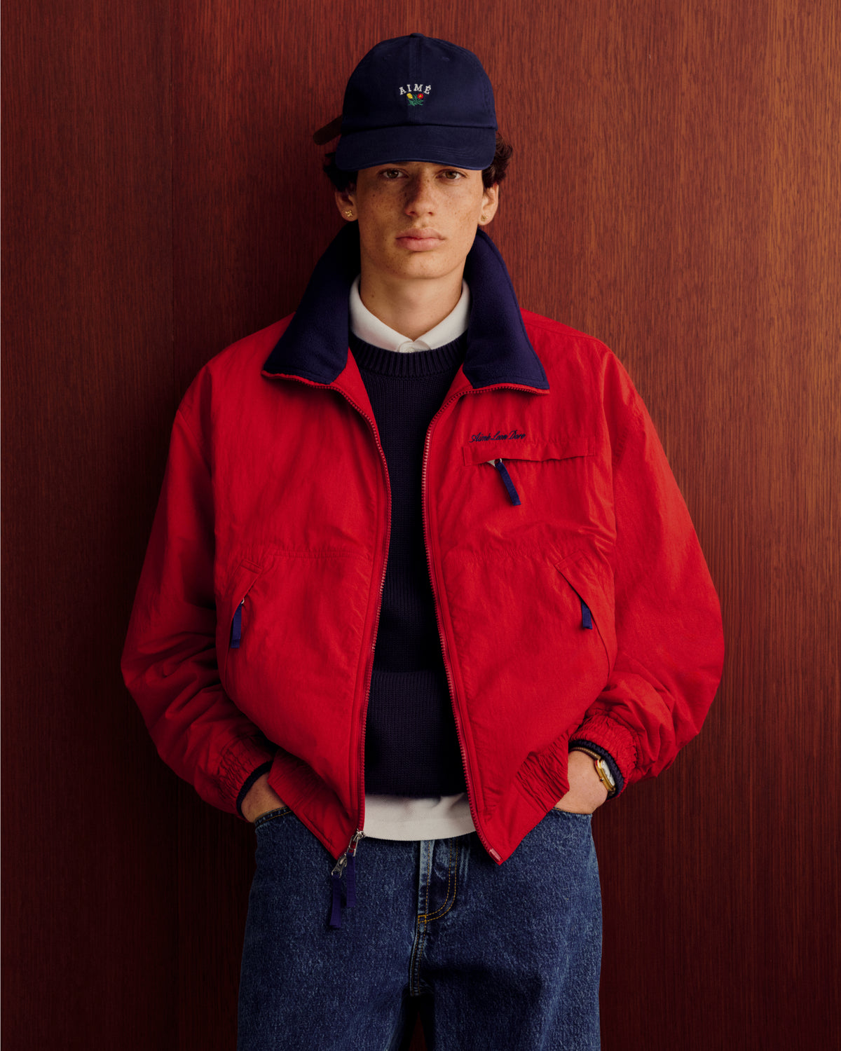 Young man in a Fleece Lined Sport Windbreaker, navy cap, and jeans stands against a wooden background, hands in pockets.