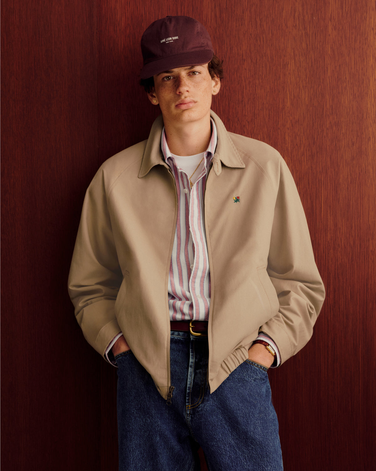 Young man in a Striped Crest Oxford Shirt, tan jacket, jeans, and burgundy cap stands by a wood-paneled wall.