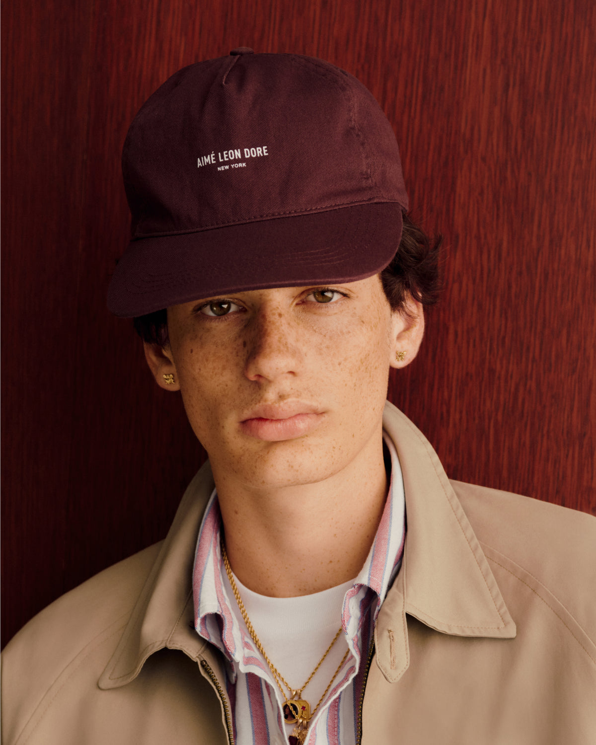 Young person in a Micro Logo Hat, beige jacket, and striped shirt stands before a red textured background.