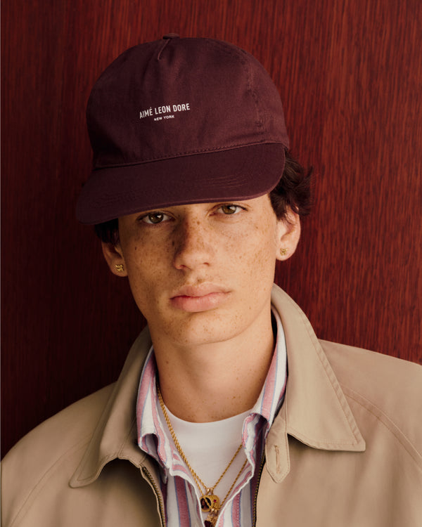 Young person in a Micro Logo Hat, beige jacket, and striped shirt stands before a red textured background.