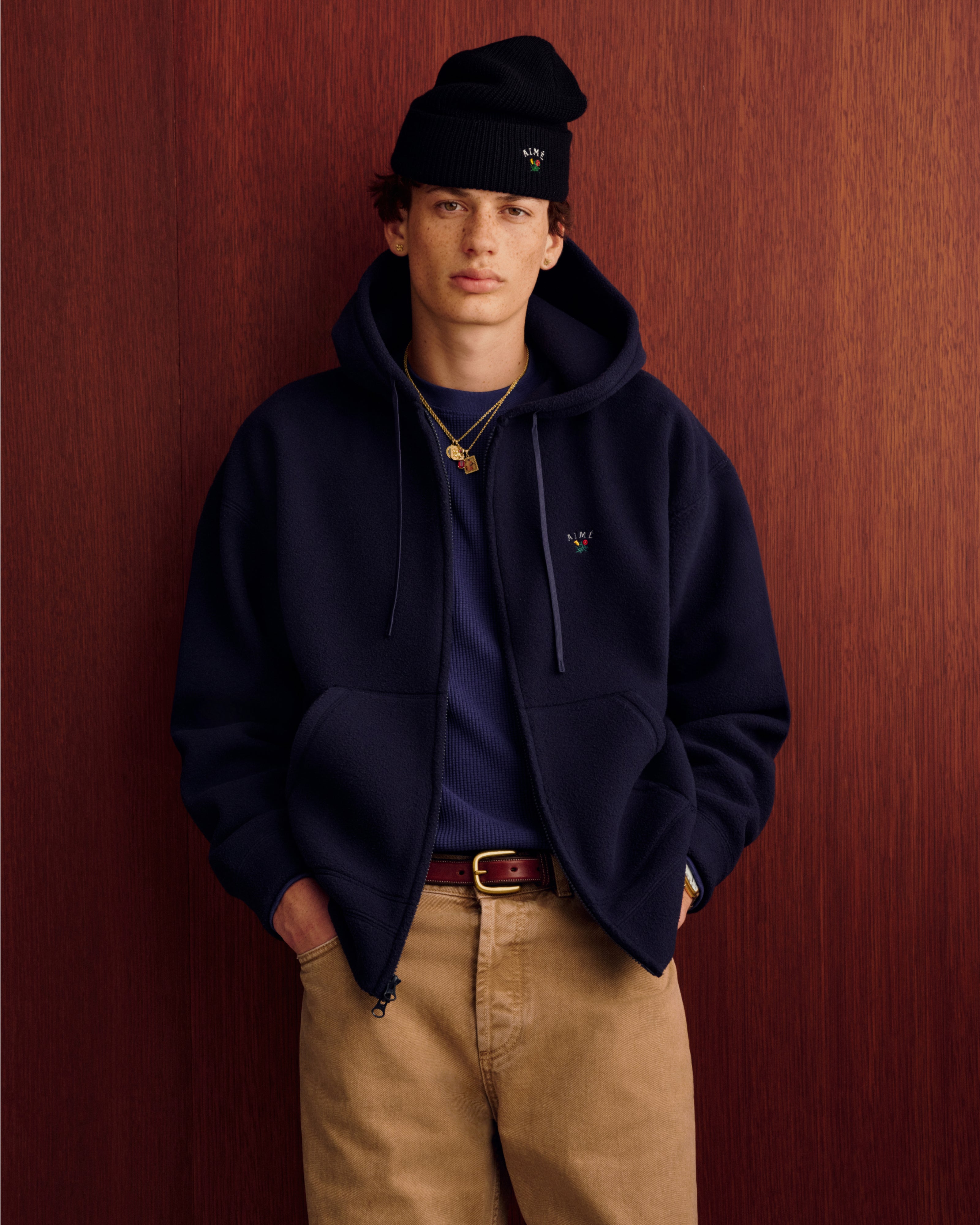 Young person in a Crest Fleece Full-Zip Hoodie, navy shirt, khakis, and black beanie by a wood paneled wall.