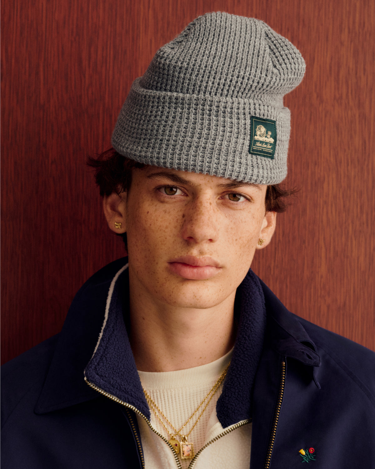 Freckled youth in gray Waffle Knit Beanie, gold jewelry, cream shirt, navy jacket, by wood panel.