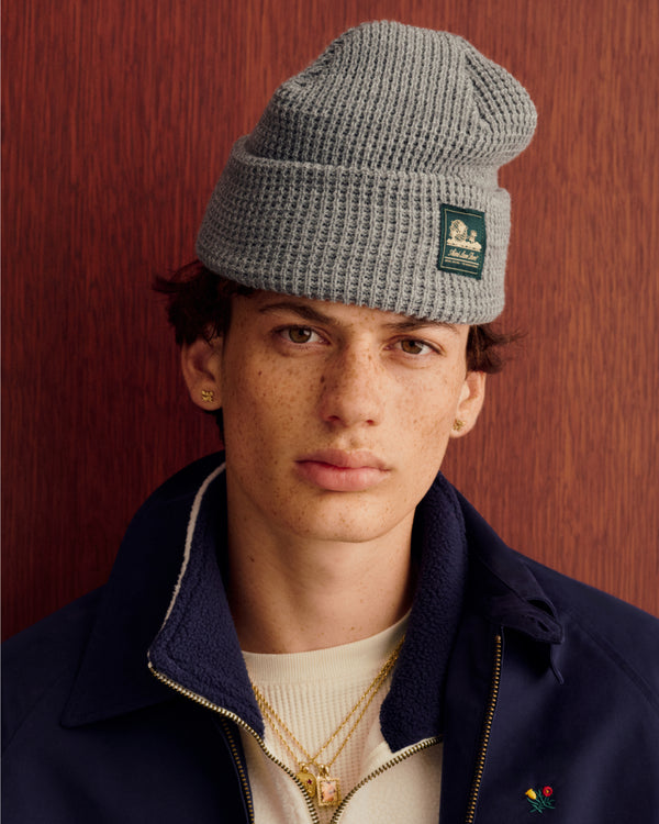 Freckled youth in gray Waffle Knit Beanie, gold jewelry, cream shirt, navy jacket, by wood panel.