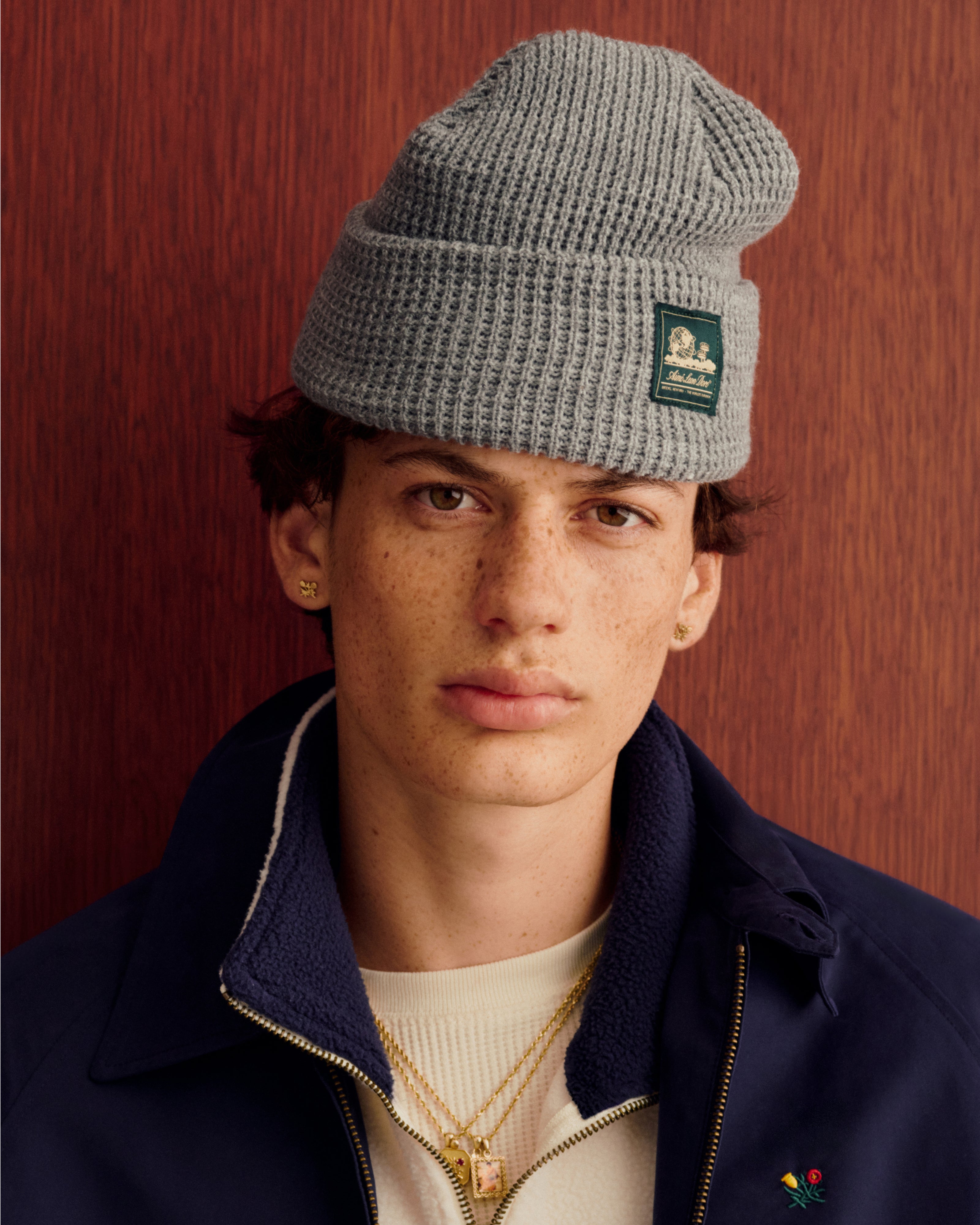 Young person in Crest Casual Jacket and gray knit beanie, with gold jewelry, against a wooden background.