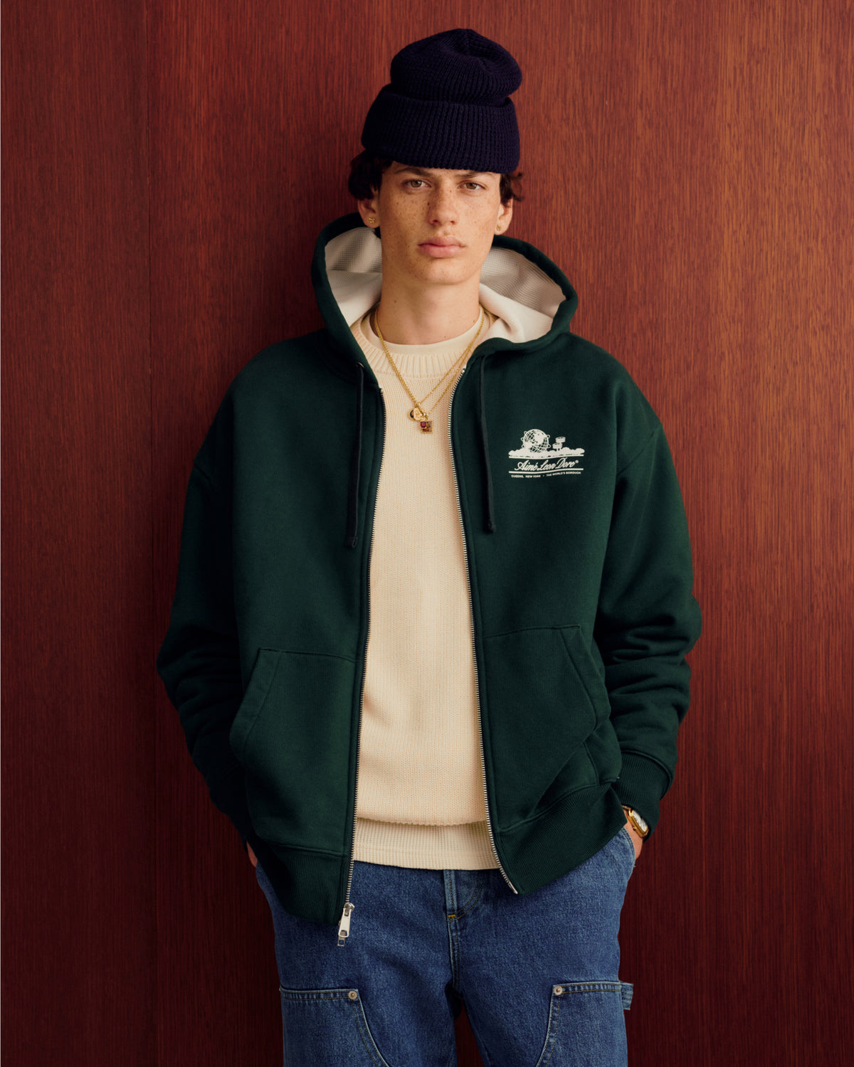 Young man in green hoodie, Crest Raglan Sweater, blue jeans, and navy beanie stands by a wooden background.