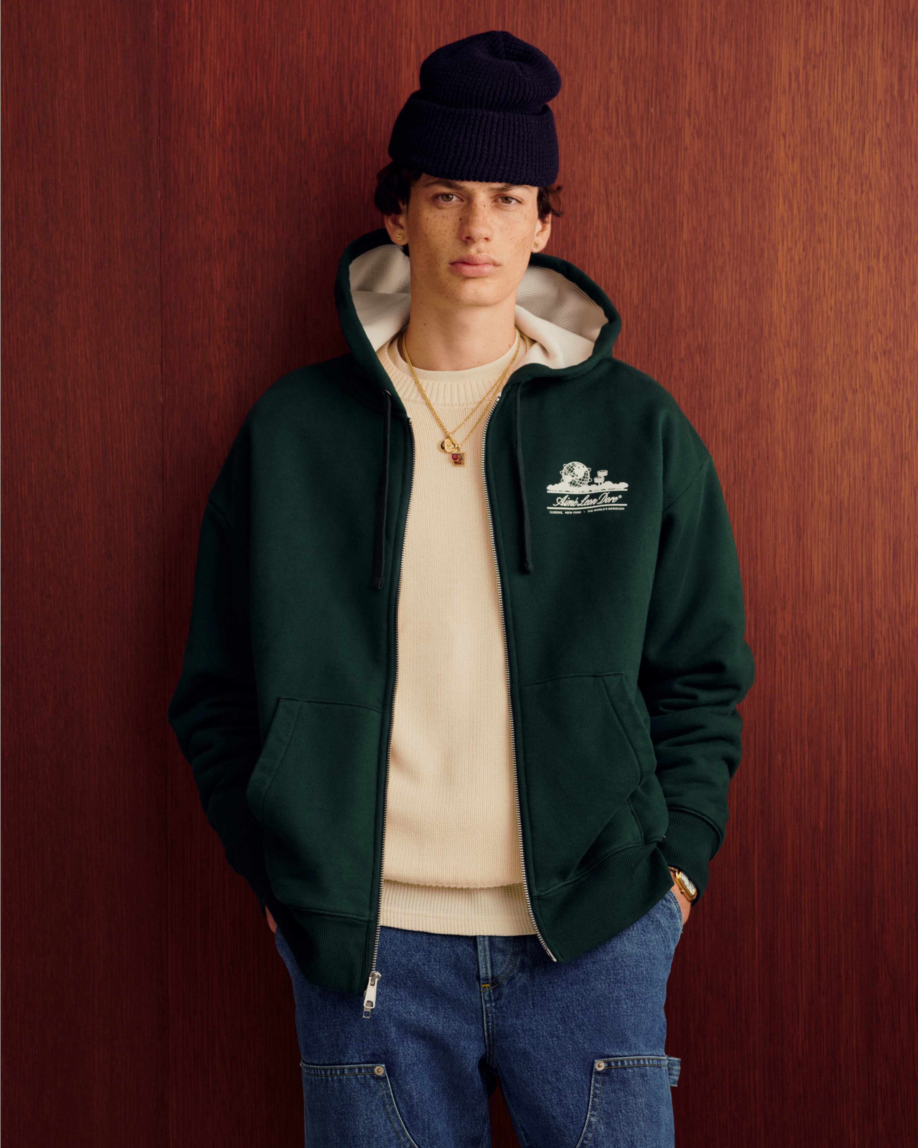 Young man in green hoodie, Crest Raglan Sweater, blue jeans, and navy beanie stands by a wooden background.