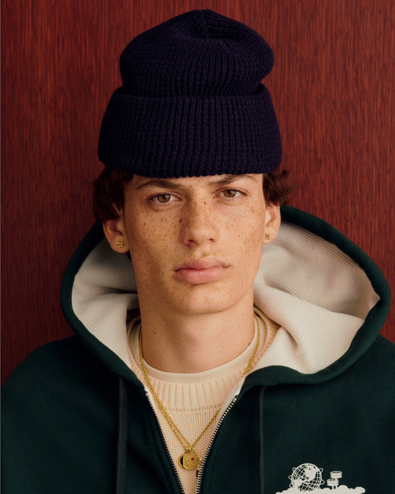 Young person in a navy Waffle Knit Beanie, green hoodie, and gold necklaces stands by a wood background.