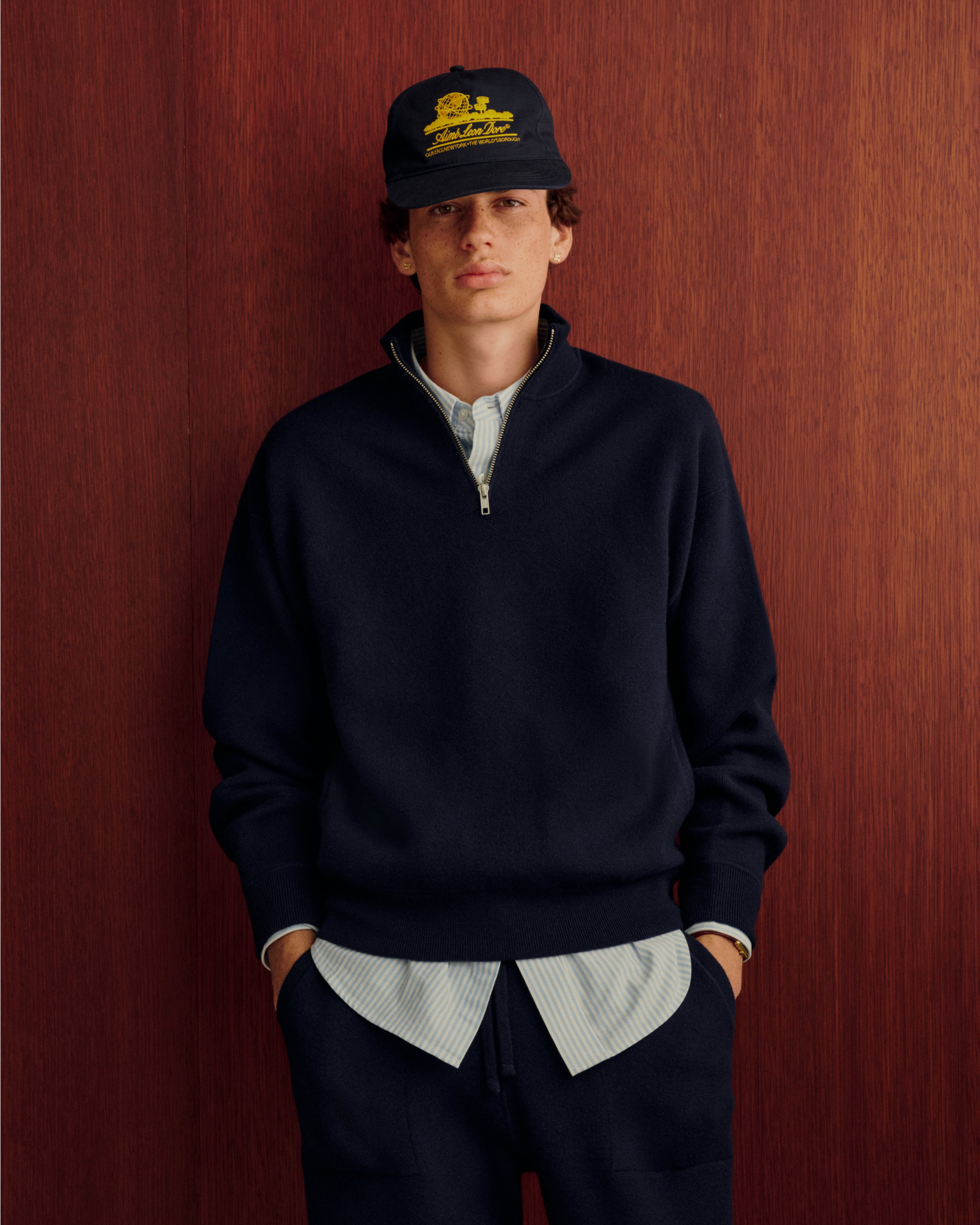 A young man in Pavilion Cashmere Sweatpants and a dark cap stands against wood, hands in pockets.