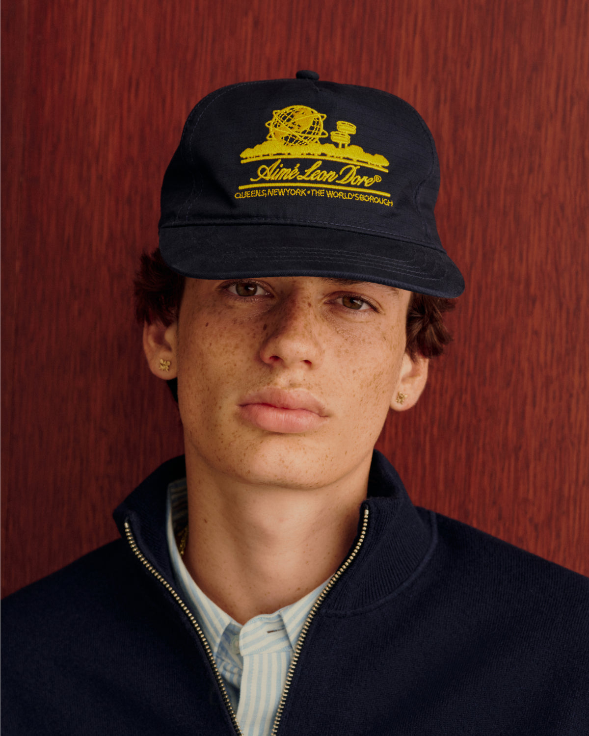 Young person in a Unisphere Hat, navy jacket, and striped shirt, standing by a wood background.