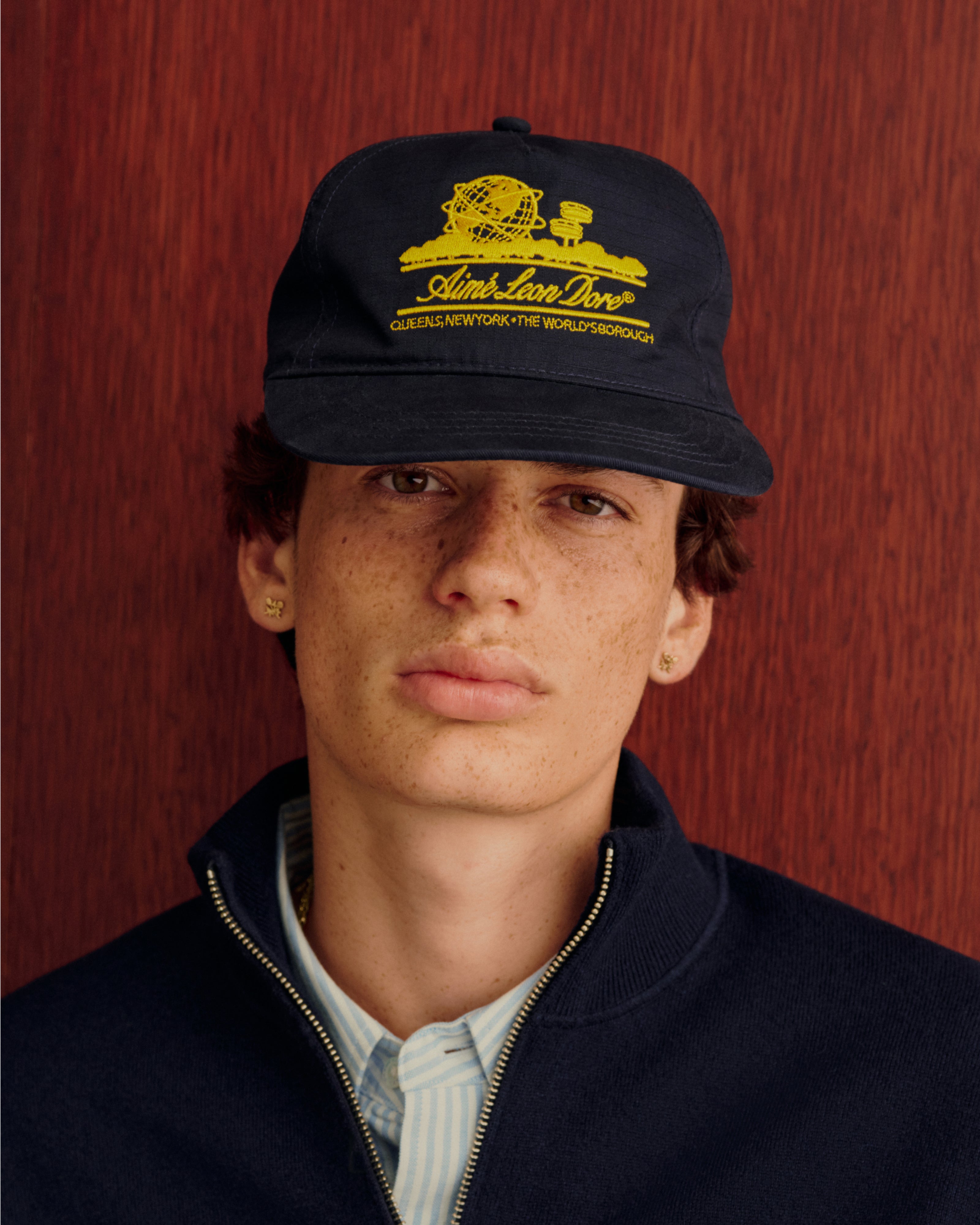 Young person in a Unisphere Hat, navy jacket, and striped shirt, standing by a wood background.