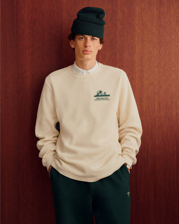 Man in cream sweater, white shirt, green pants, and Waffle Knit Beanie stands by wood wall, hands in pockets.
