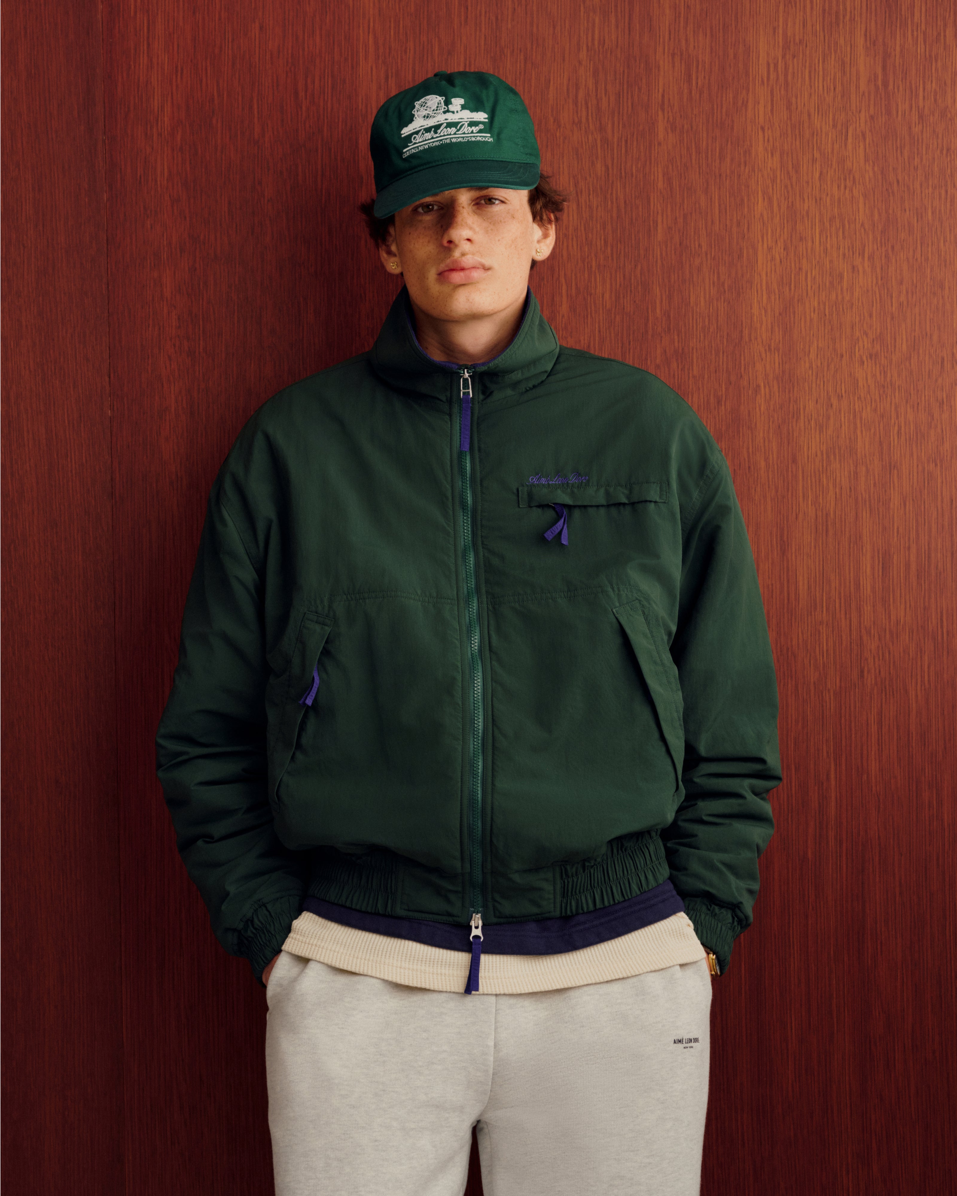 Individual wearing a green Fleece Lined Sport Windbreaker, cream sweatshirt, and gray pants stands by a wooden wall.