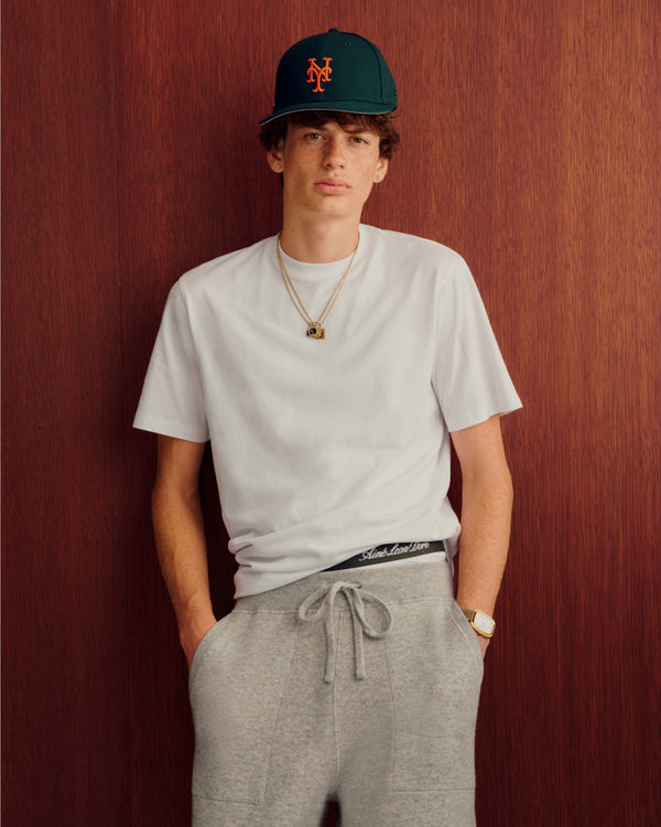 Young man in a white tee, gray sweats, and ALD/New Era Mets Ballpark Hat stands before a wood wall.