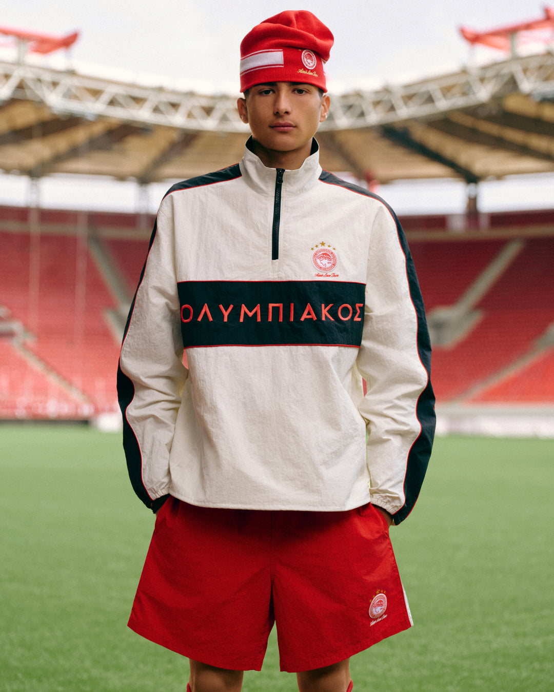 A person wearing an ALD / Olympiacos F.C. Nylon Quarter Zip Pullover and a red beanie on a soccer field.