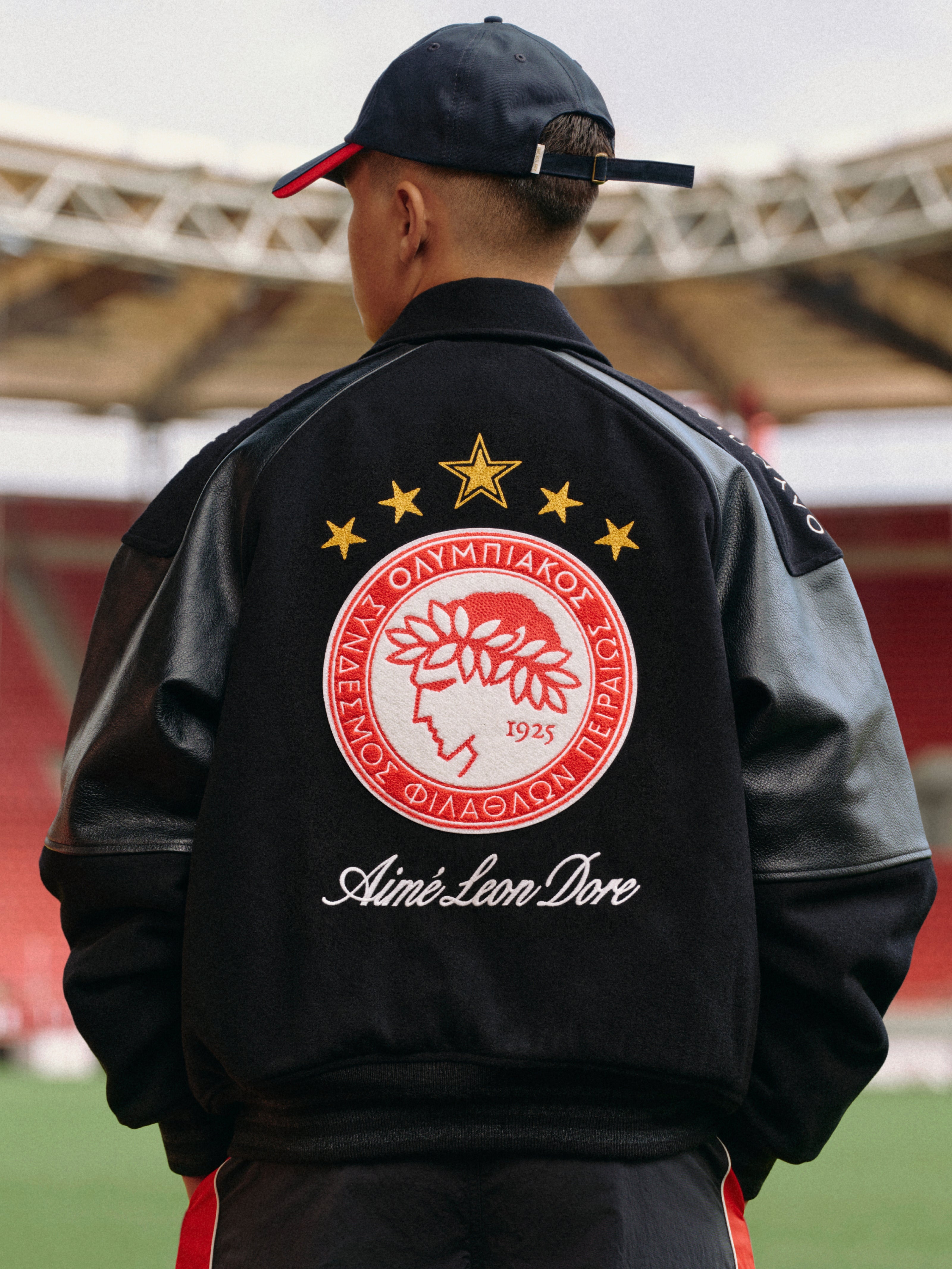 Person wearing an ALD / Olympiacos F.C. Team Leather Jacket with a red and white club emblem on the back.