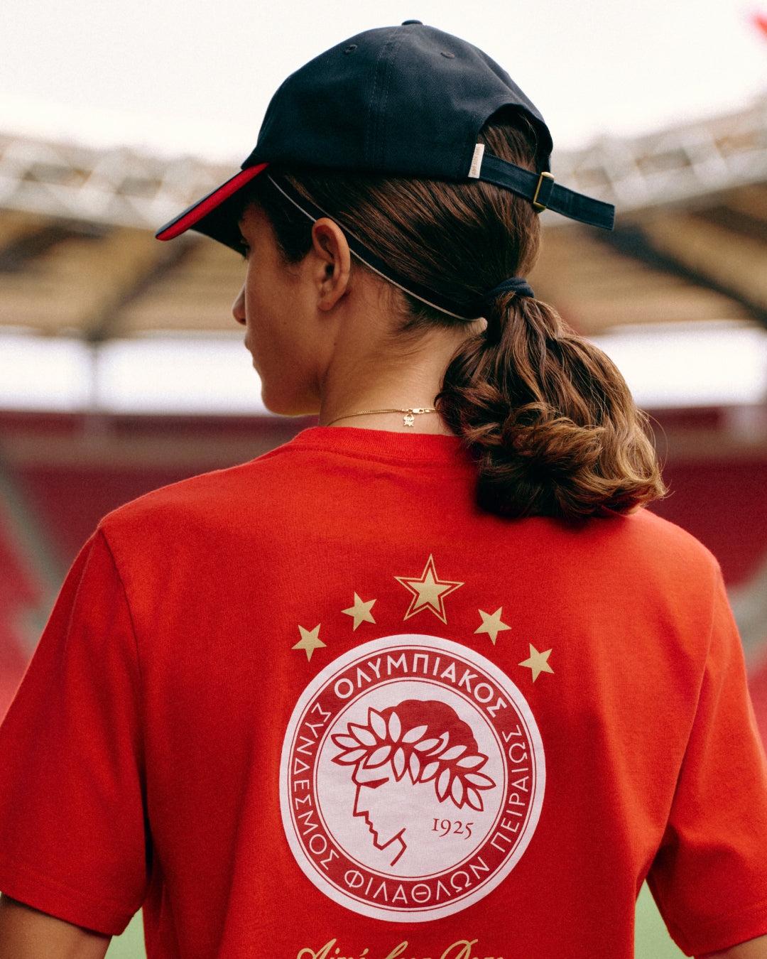 A person wears a red ALD / Olympiacos F.C. Souvenir Tee with a laurel-wreathed profile logo and five stars above it.