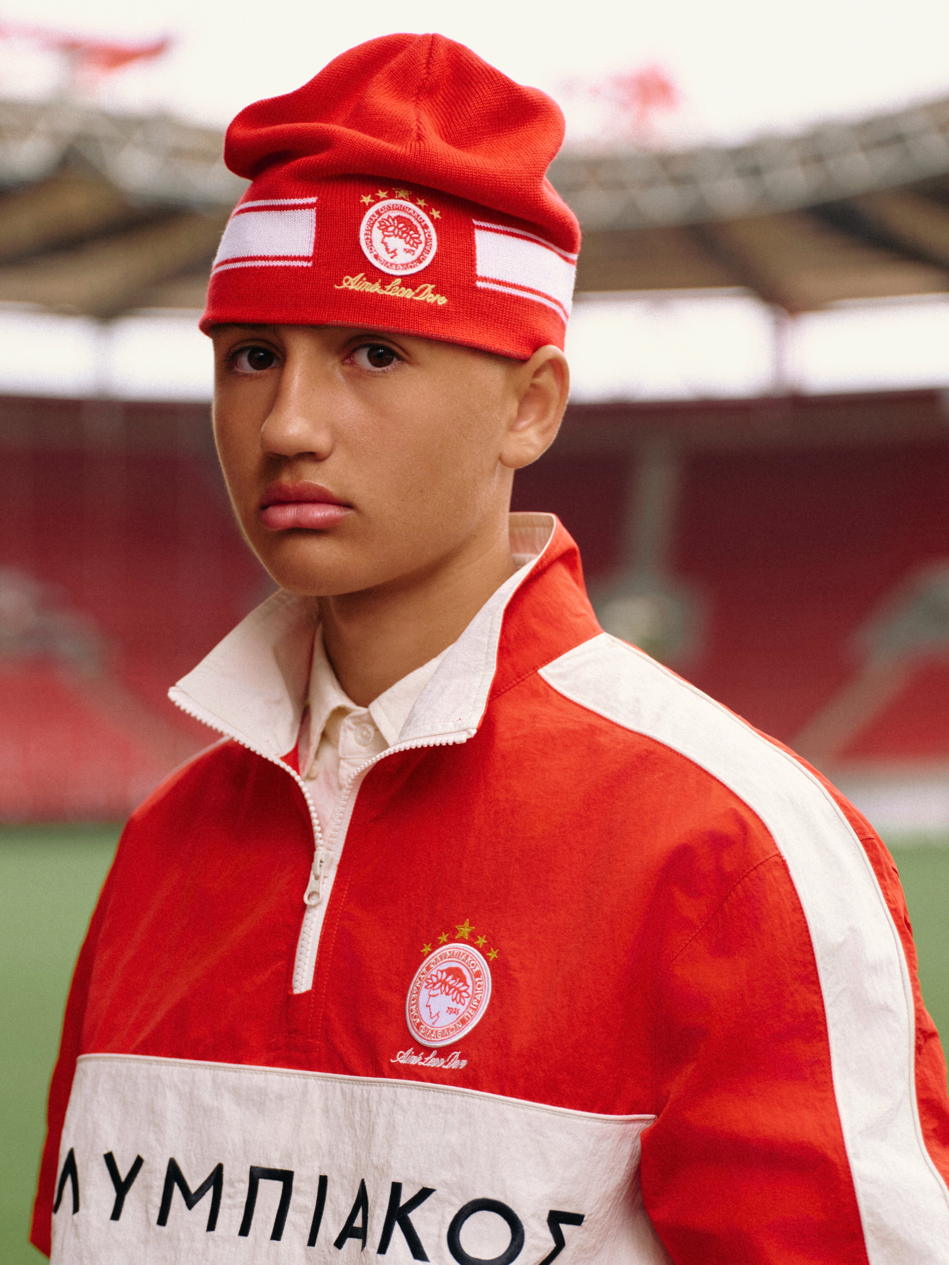 A person wearing a red beanie and an ALD / Olympiacos F.C. Nylon Quarter Zip Pullover in a stadium.