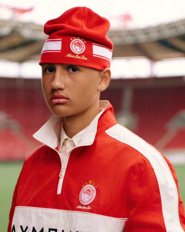 A person wearing an ALD / Olympiacos F.C. Stadium Beanie and a matching red and white quarter-zip jacket in a stadium.