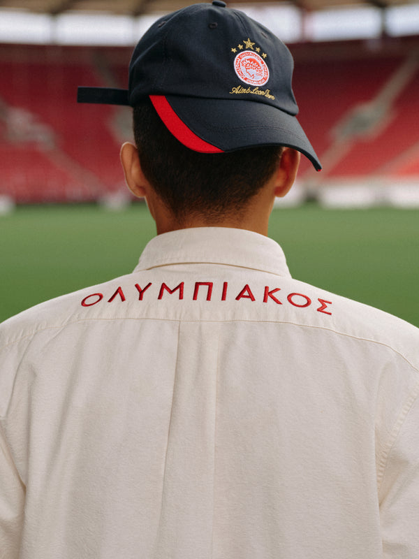 A person wearing an ALD / Olympiacos F.C. Souvenir Hat and a cream button-down shirt with Greek lettering on the shoulders.