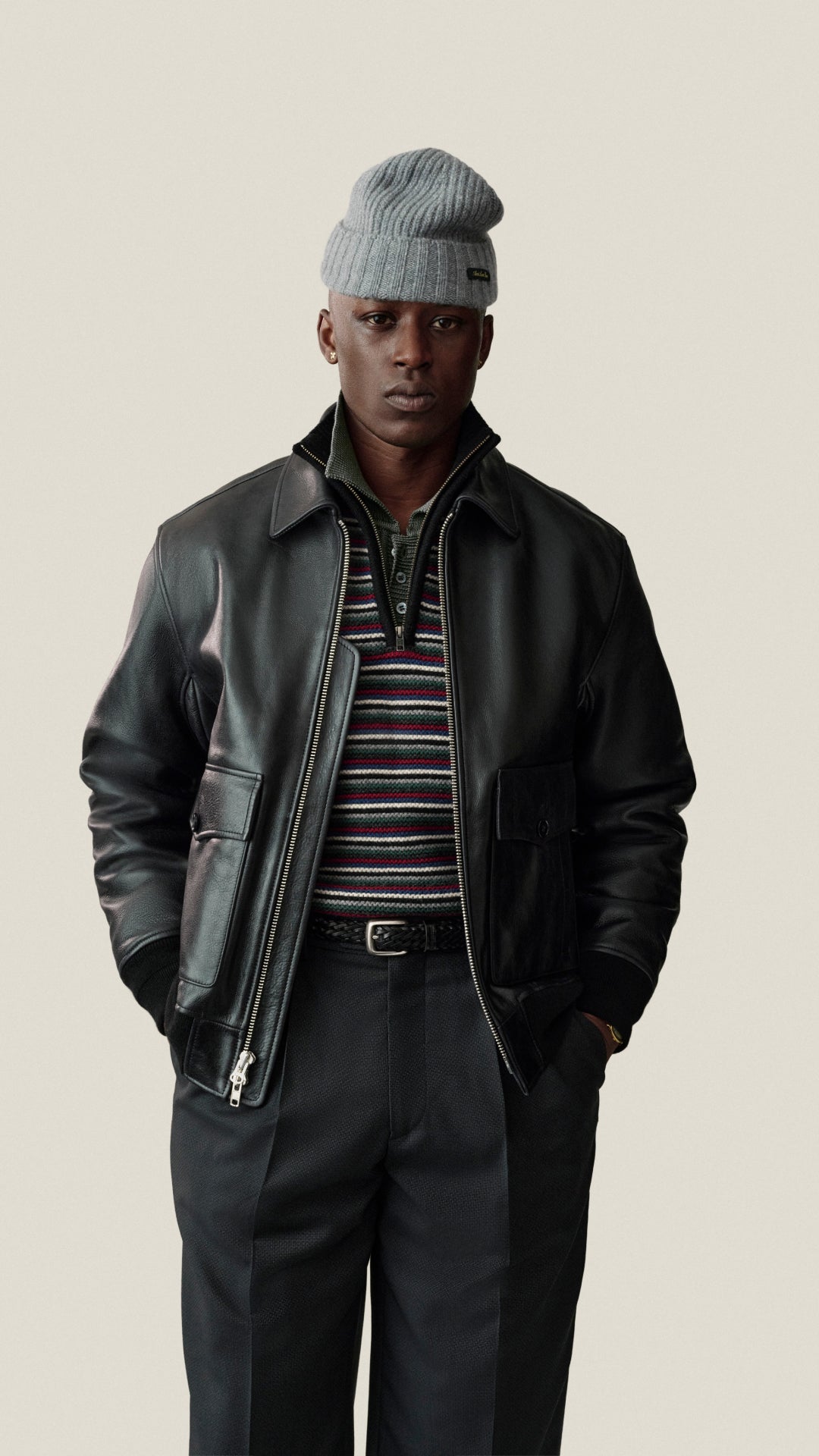 A man wearing a black leather jacket, a gray knit beanie, a striped polo shirt, and dark trousers, standing against a solid light beige background.