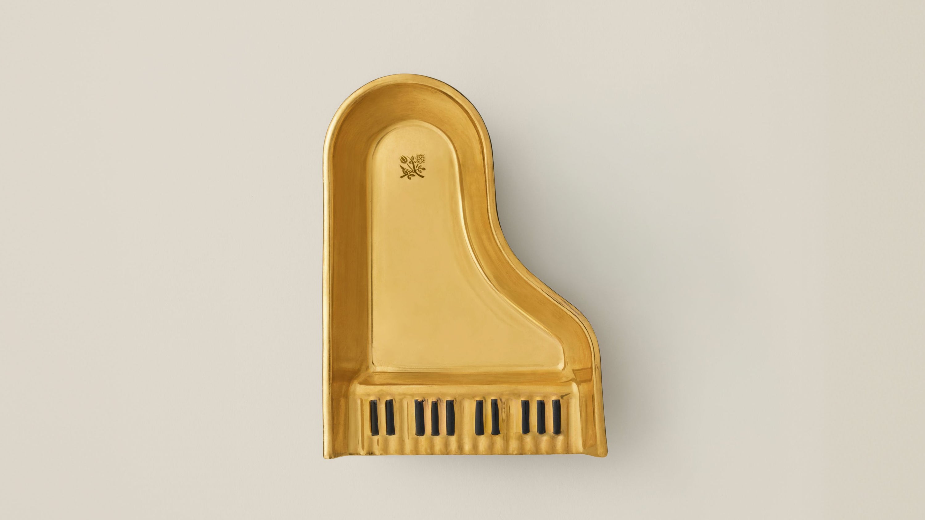 A gold-finished ceramic grand piano ashtray featuring black key accents at the bottom, shown against a neutral light beige background.