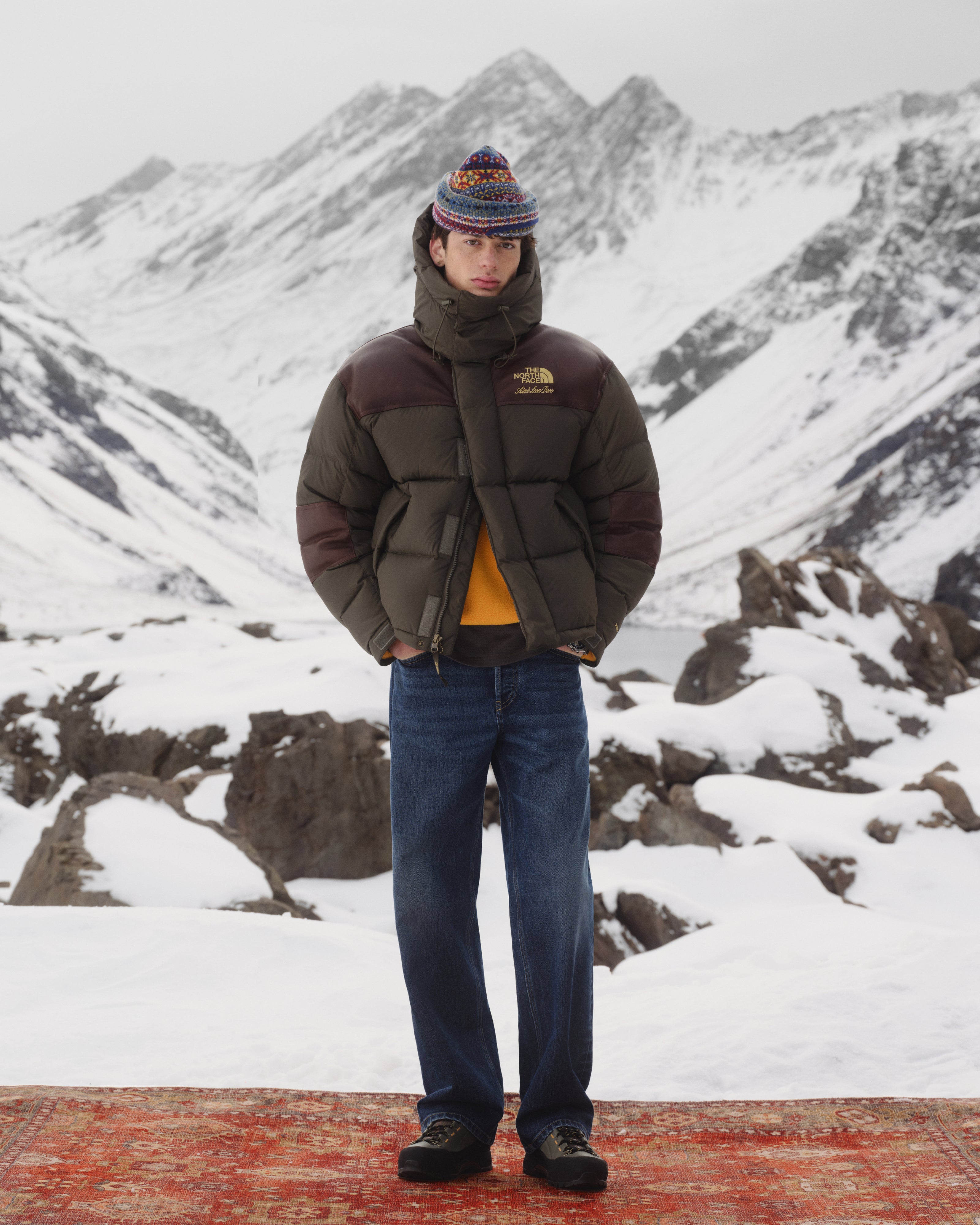Person in ALD / The North Face Baltoro Jacket, jeans, and knit hat stands on a rug in snowy mountains.