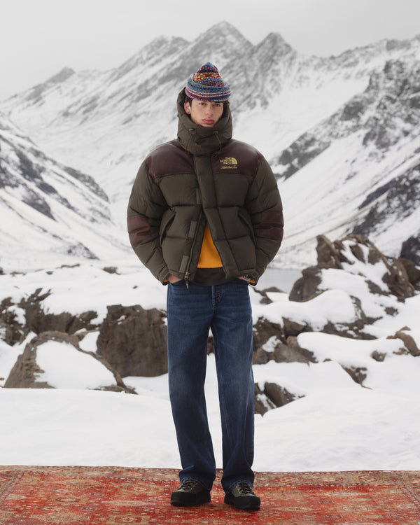 Person in ALD / The North Face Baltoro Jacket, jeans, and knit hat stands on a rug in snowy mountains.