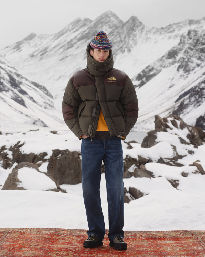Person in ALD / The North Face Baltoro Jacket, jeans, and knit hat stands on a rug in snowy mountains.