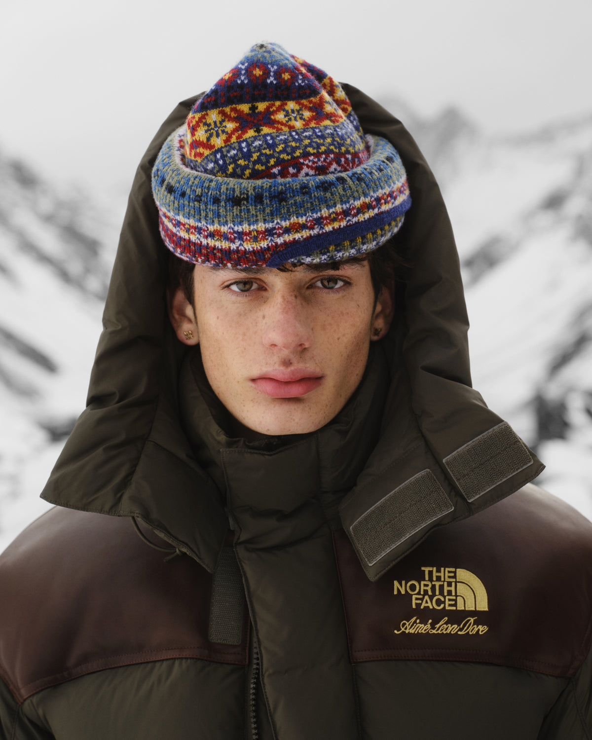 Young person in an ALD / The North Face Baltoro Jacket and knit hat stands in snowy mountains, facing the camera.